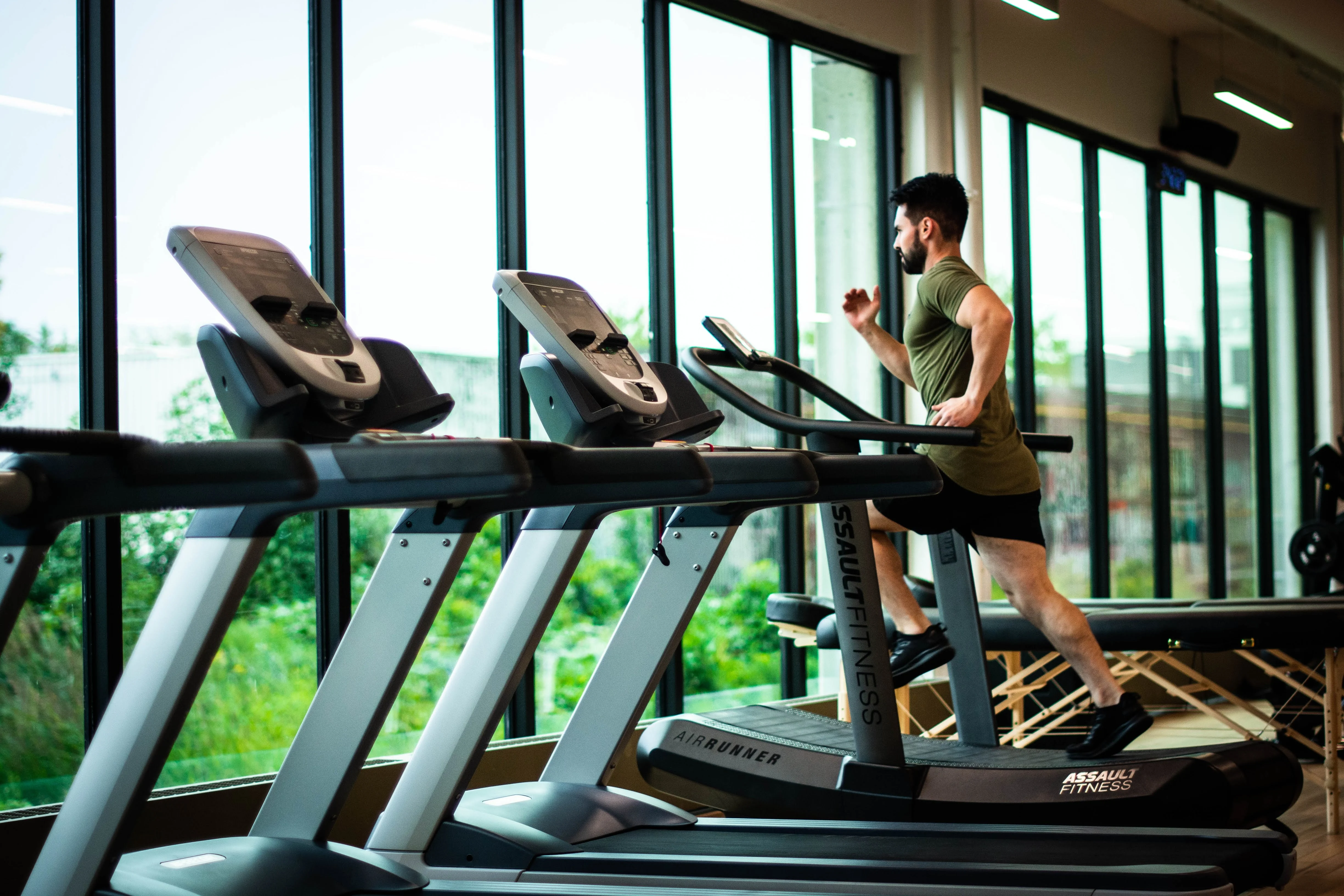 A man running on a treadmill in a modern gym with large windows showing greenery outside.