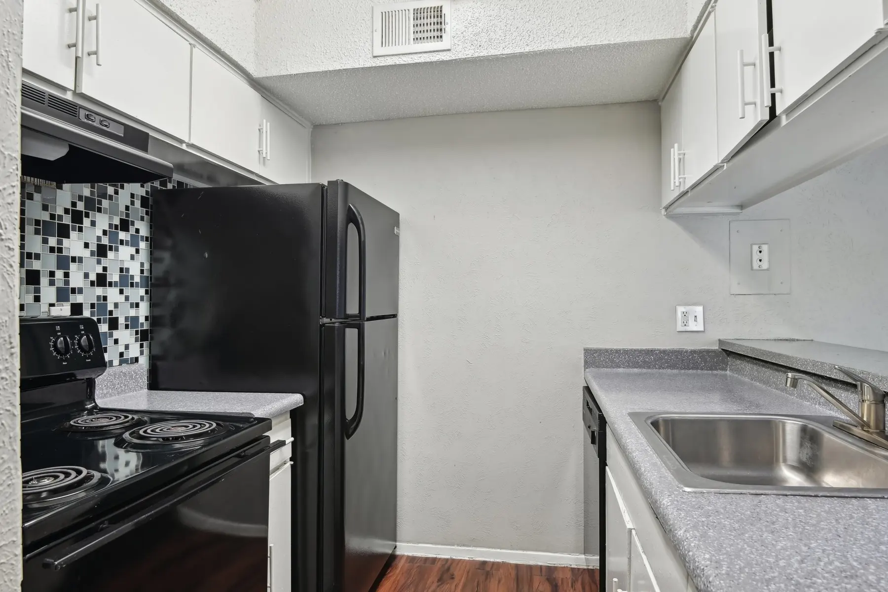 A compact kitchen featuring a black stove, refrigerator, and two-toned backsplash. The countertops are gray with a stainless steel sink and white cabinetry.
