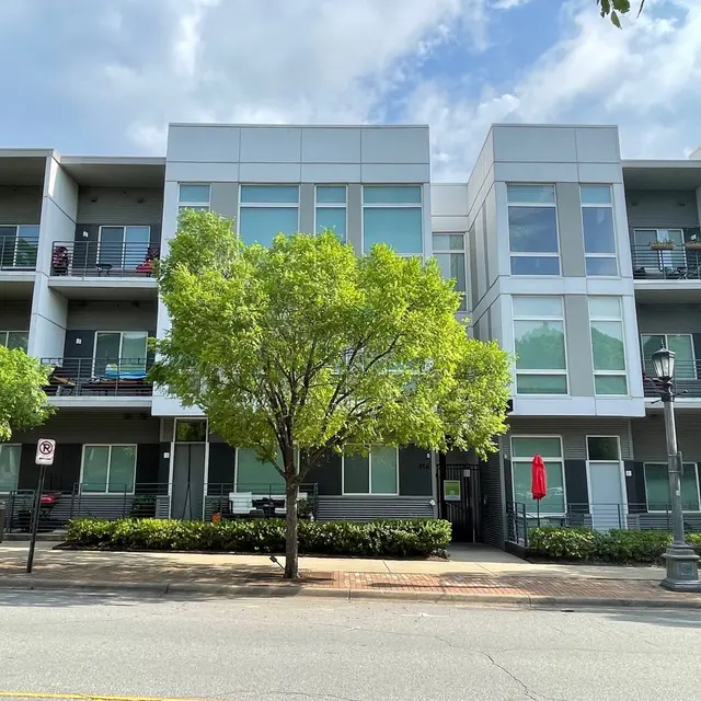 A modern apartment building with multiple stories, featuring balconies and large windows, surrounded by green trees and a pedestrian path.