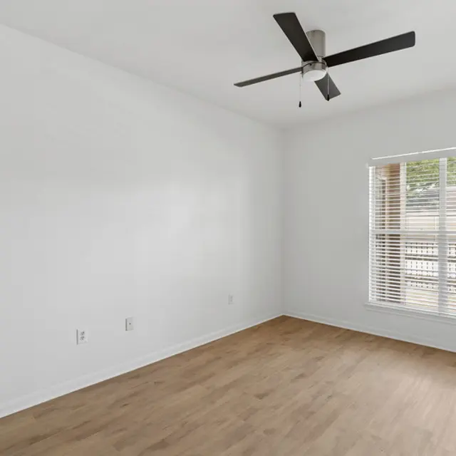 An empty room with light-colored wooden flooring, a ceiling fan, and a window with blinds that allow light to enter. The walls are painted white, enhancing the room's brightness and spaciousness. A closed door is on the left side.