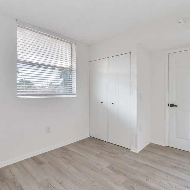 A bright and empty room with white walls, a window with blinds, and wooden flooring. There is a closet with sliding doors on one side and a door leading out on the opposite side.