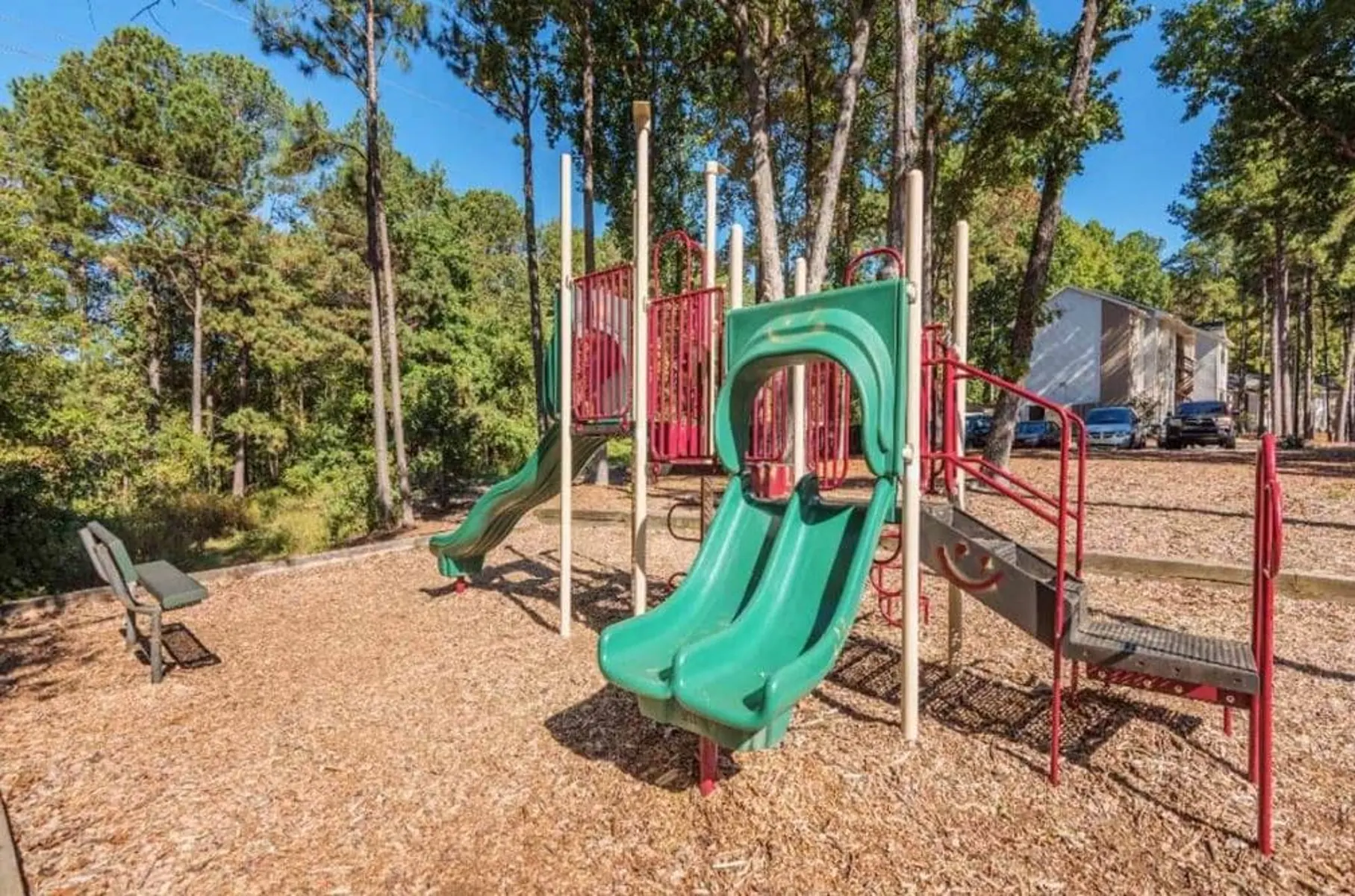A playground featuring two green slides, surrounded by trees and wooded area with wood chip ground cover. A bench is visible in the foreground.