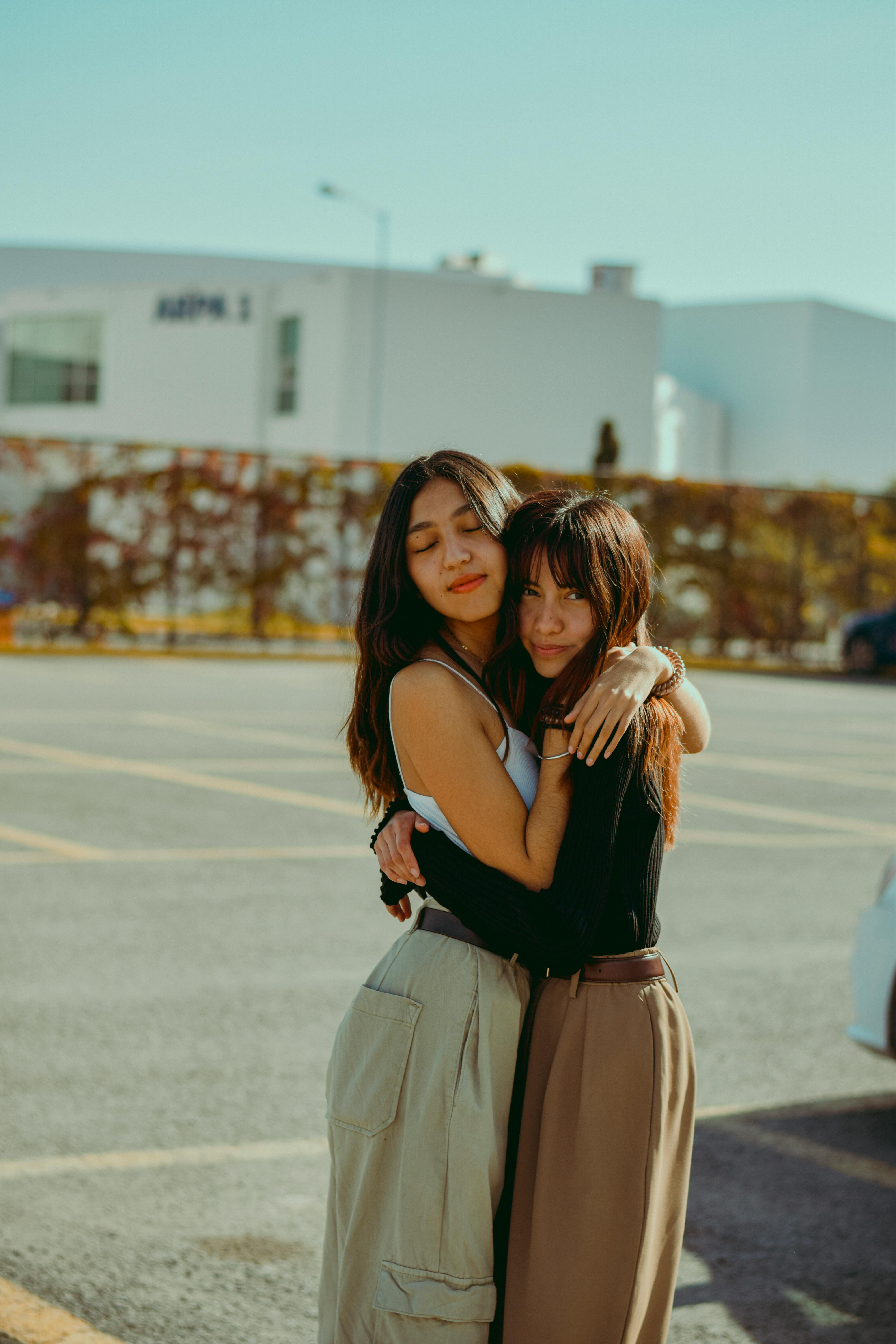 Friends Embracing Two young women embracing outdoors, smiling at the camera; one wearing a black top and the other wearing a white tank top with a long light skirt.