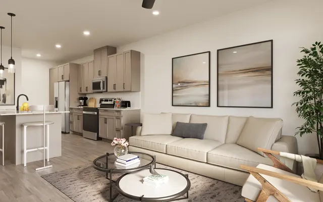 A modern living room featuring a light-colored sectional sofa, a coffee table, and a chair. The kitchen is in the background with light wood cabinetry and stainless steel appliances. There are two abstract art pieces on the wall and a potted plant in the corner.