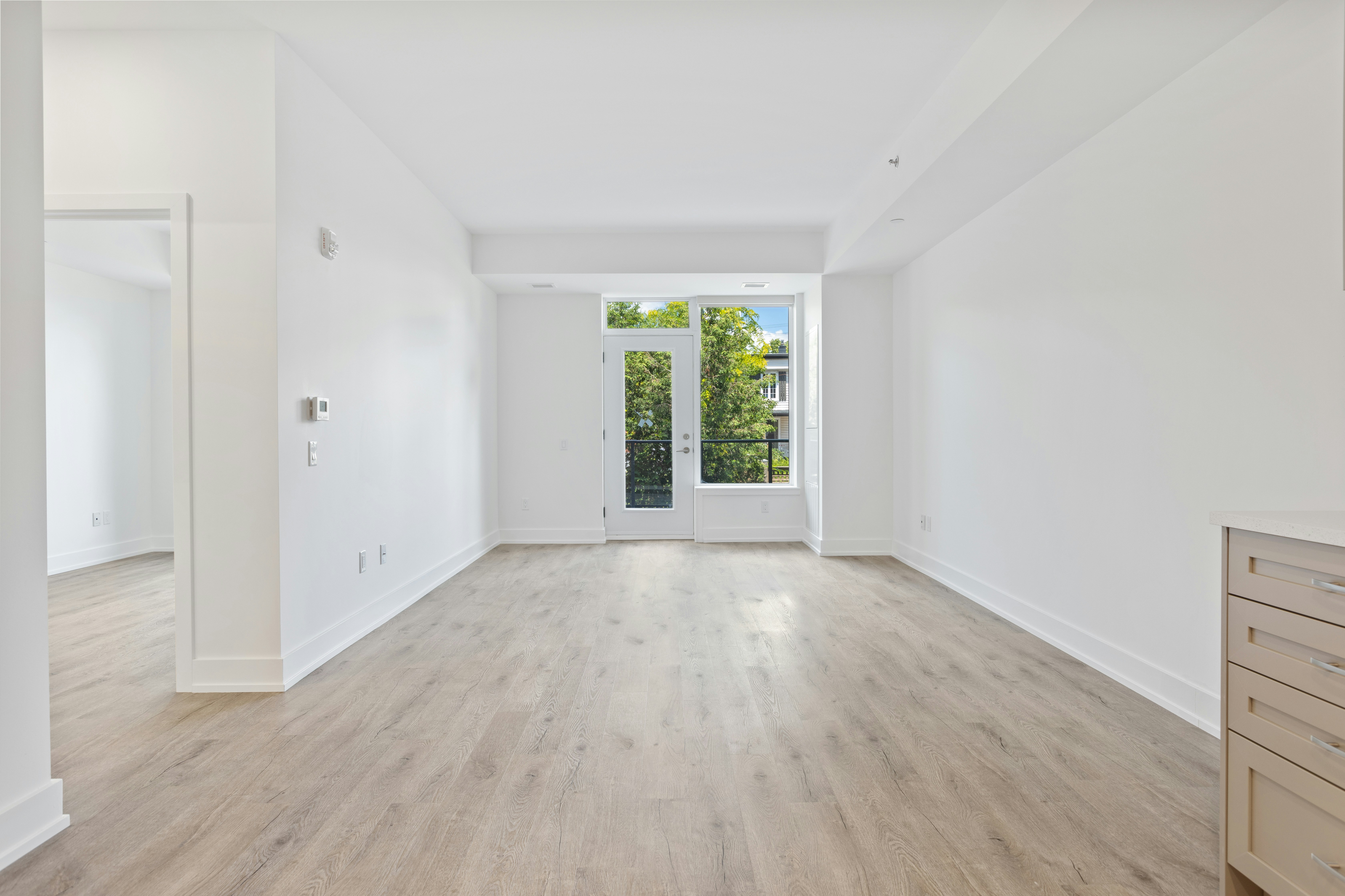 Bright and airy living room with large windows and light wood flooring, featuring white walls and a view of greenery outside.