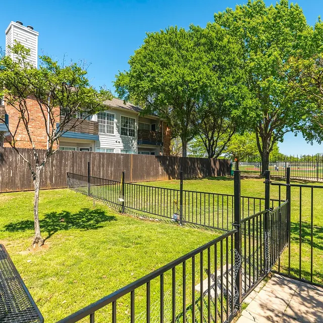 A fenced area of a backyard with open grass space, trees, and a bench on the side. In the background, there are residential buildings.