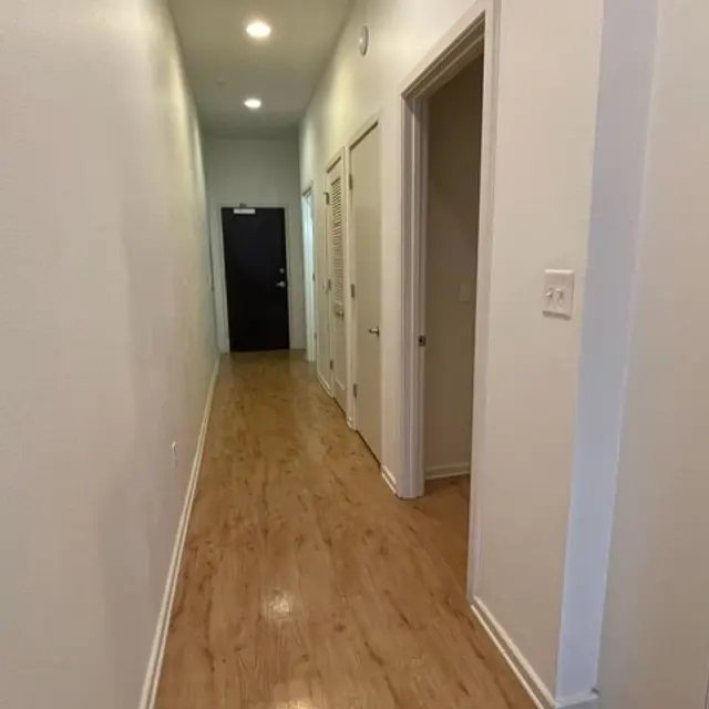 A view of a long hallway featuring light-colored walls and wooden flooring. The hallway has several doors on the right side and a dark door at the end.