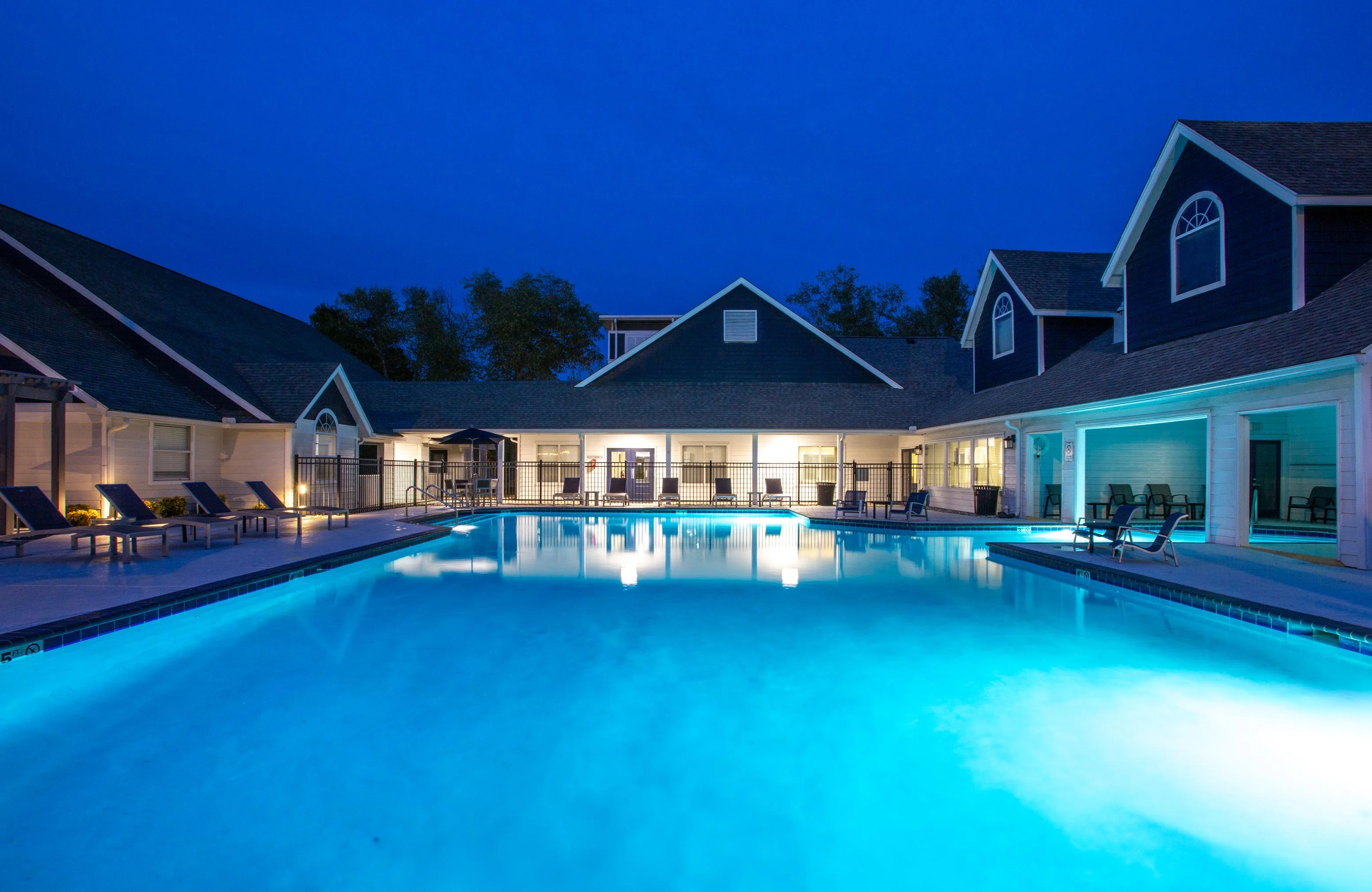 A brightly lit outdoor pool area at night, surrounded by lounge chairs and buildings with large windows.