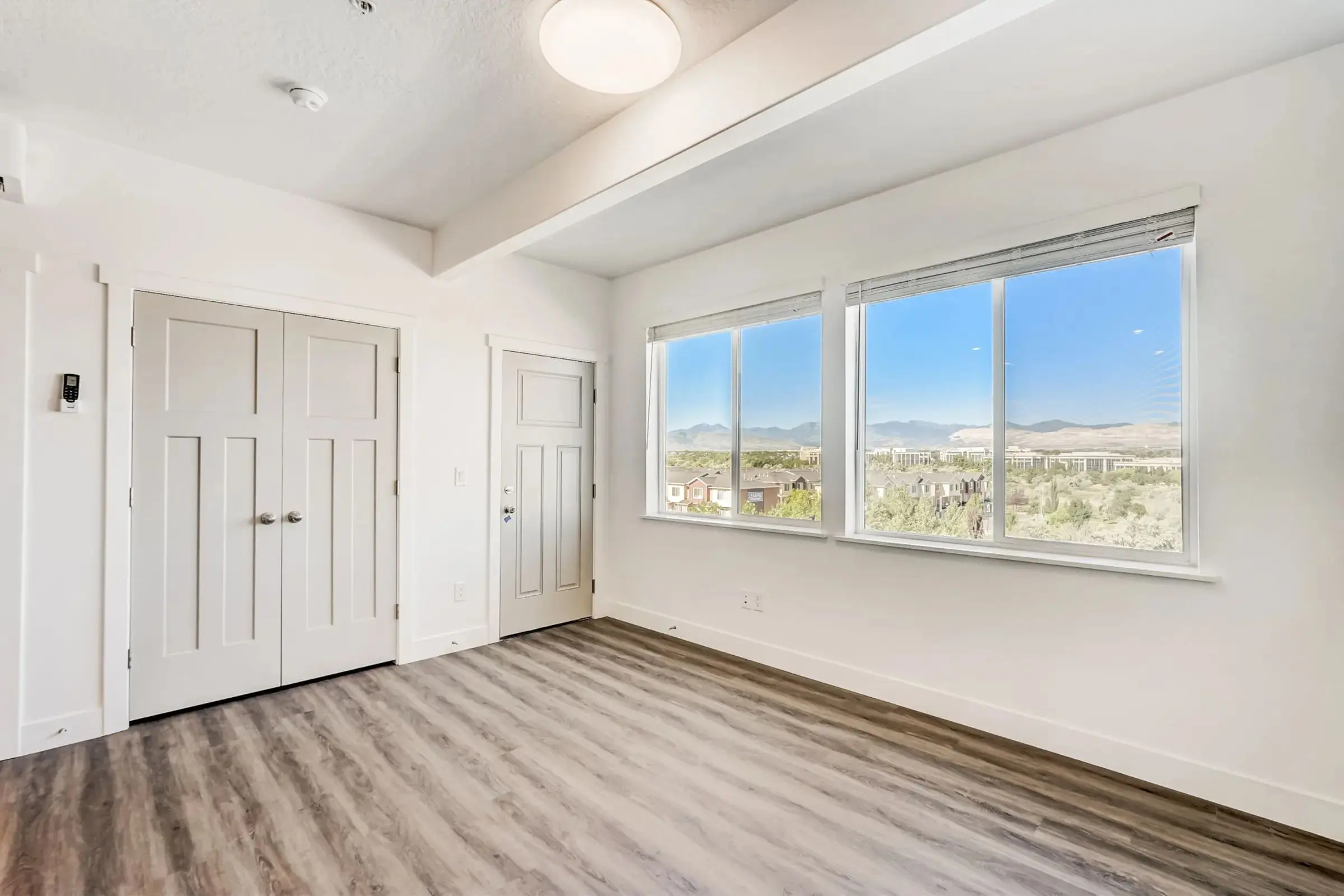 Spacious Empty Room with Scenic View A spacious empty room with large windows and a scenic view. The room features wood-like flooring and a white ceiling with a light fixture. Two closed doors are visible on the left side of the room. The windows open to a view of hills and trees.