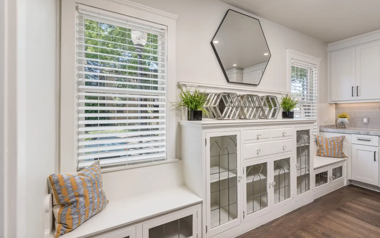 A bright kitchen nook featuring white cabinetry, built-in seating with striped pillows, and large windows with white blinds letting in natural light. A geometric mirror hangs above the cabinetry, which has glass paneled doors and plants on display.