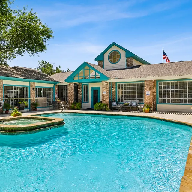 A vibrant pool area featuring a large swimming pool with a hot tub, surrounded by a stone deck and lush greenery. The building beside the pool has large windows and a welcoming design, with seating arrangements visible. It's a sunny day with a clear blue sky.