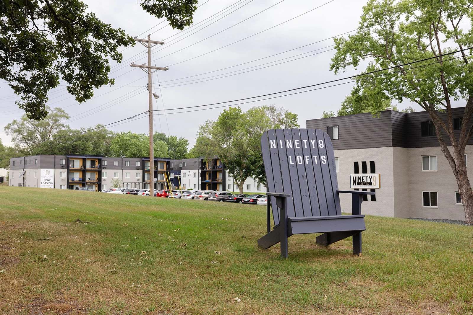 Ninety9 Lofts Apartments A large wooden chair positioned in front of an apartment complex named Ninety9 Lofts, surrounded by greenery and power lines in the background.