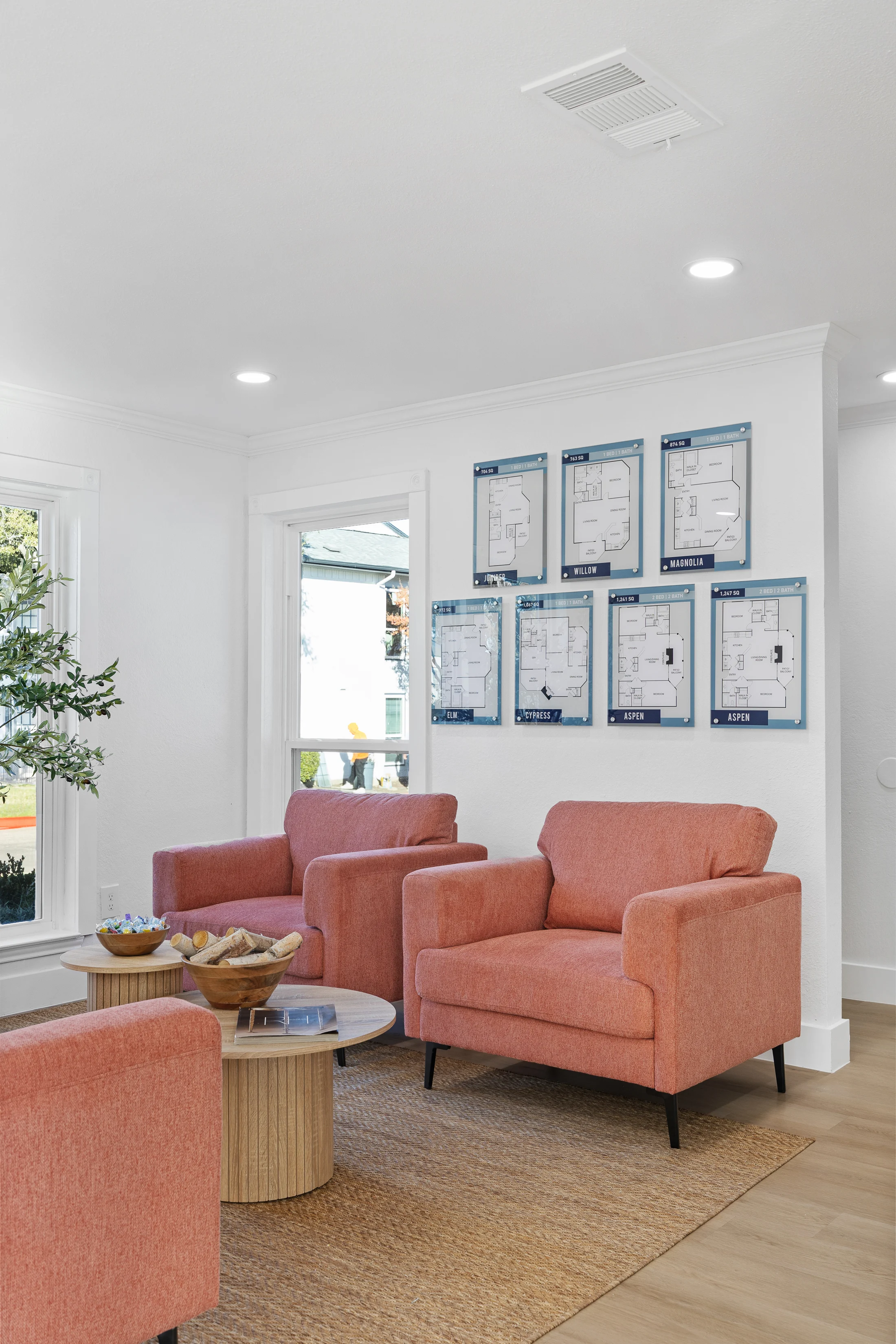 A modern living area featuring plush coral-colored chairs facing a round wooden coffee table, with a potted plant nearby and blue printouts on the wall.