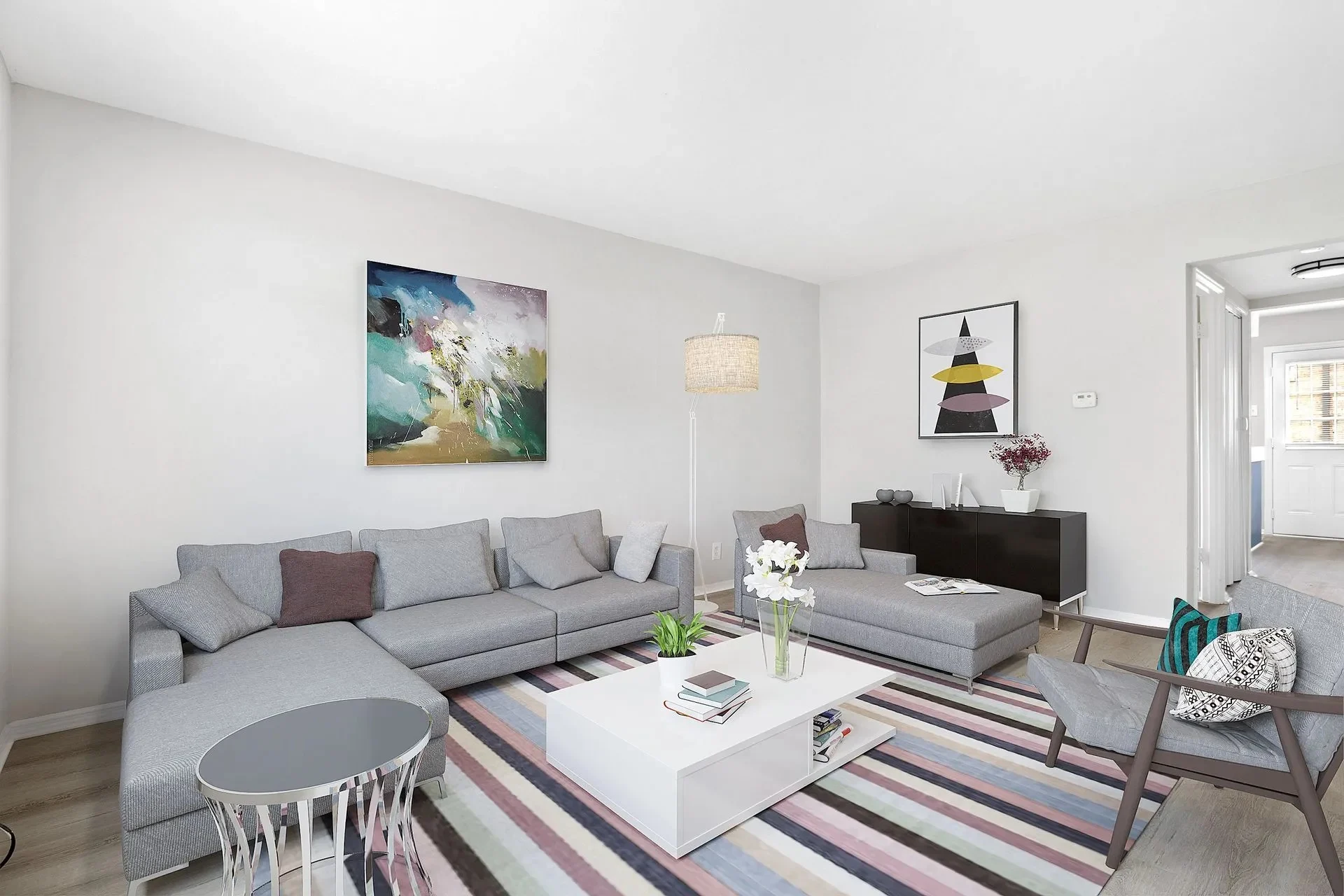 A modern living room featuring a gray sectional sofa, a round side table, and a light-colored coffee table with books and a plant on it. The walls are painted white, and there are colorful abstract paintings hanging. A sliding door leads to another room, and a floor lamp stands beside the sofa.