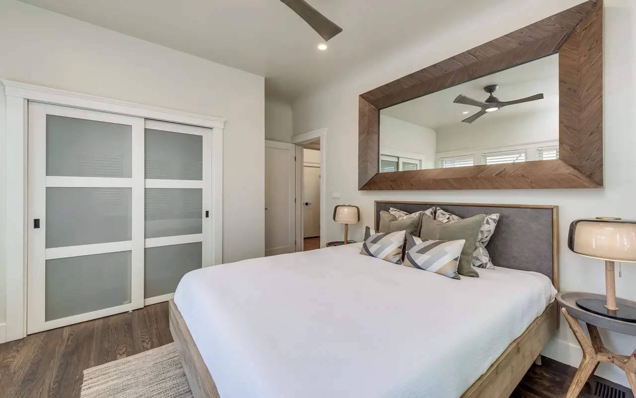 A modern bedroom featuring a queen-sized bed with crisp white linens, decorative pillows, and stylish bedside lamps. There is a large wooden framed mirror on the wall and sliding glass doors to a closet.
