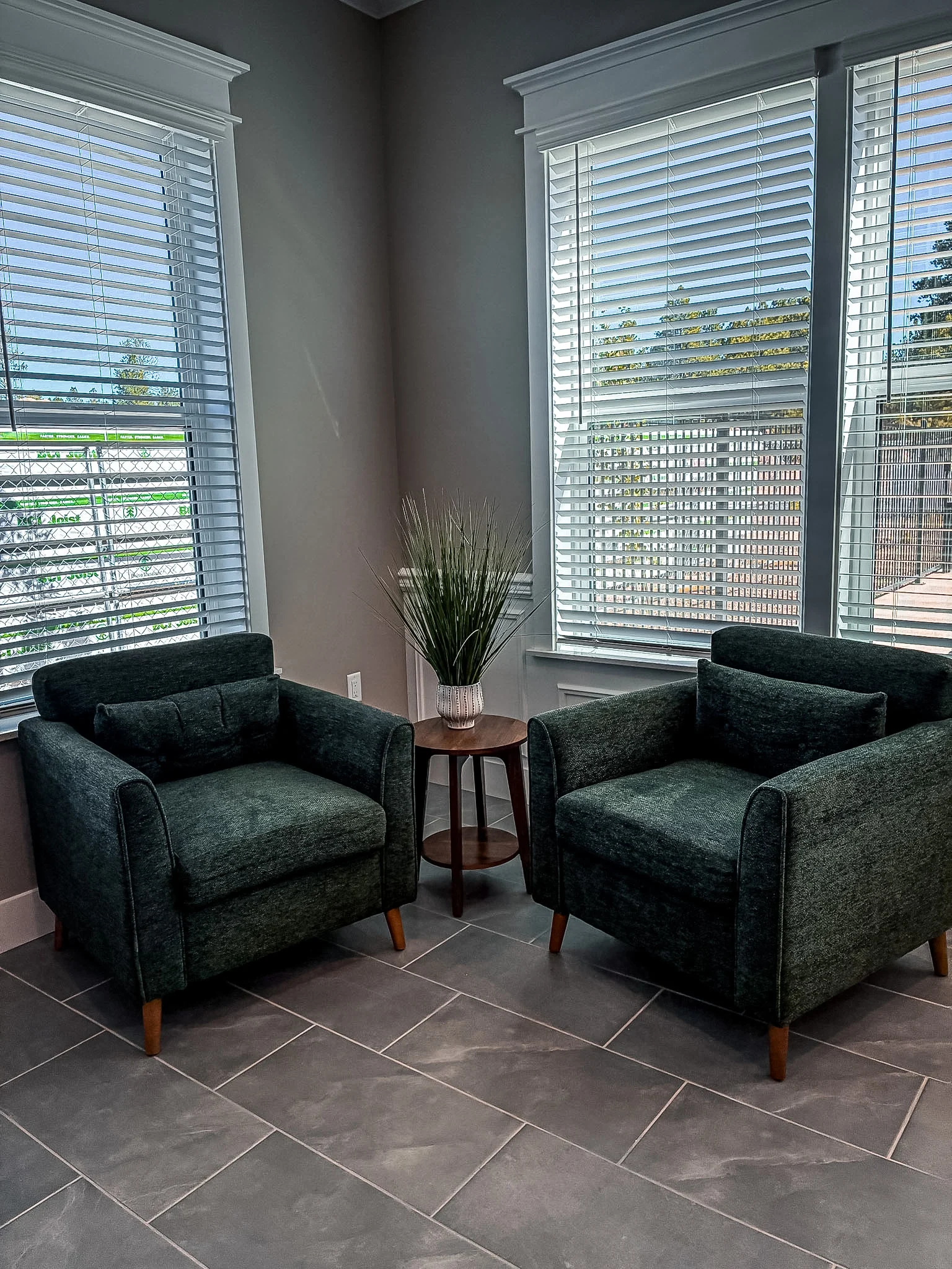 Two green armchairs in a well-lit corner with large windows and a small table between them, decorated with a plant.