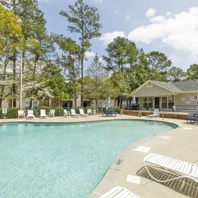 A swimming pool surrounded by lounge chairs, with trees and a building in the background.