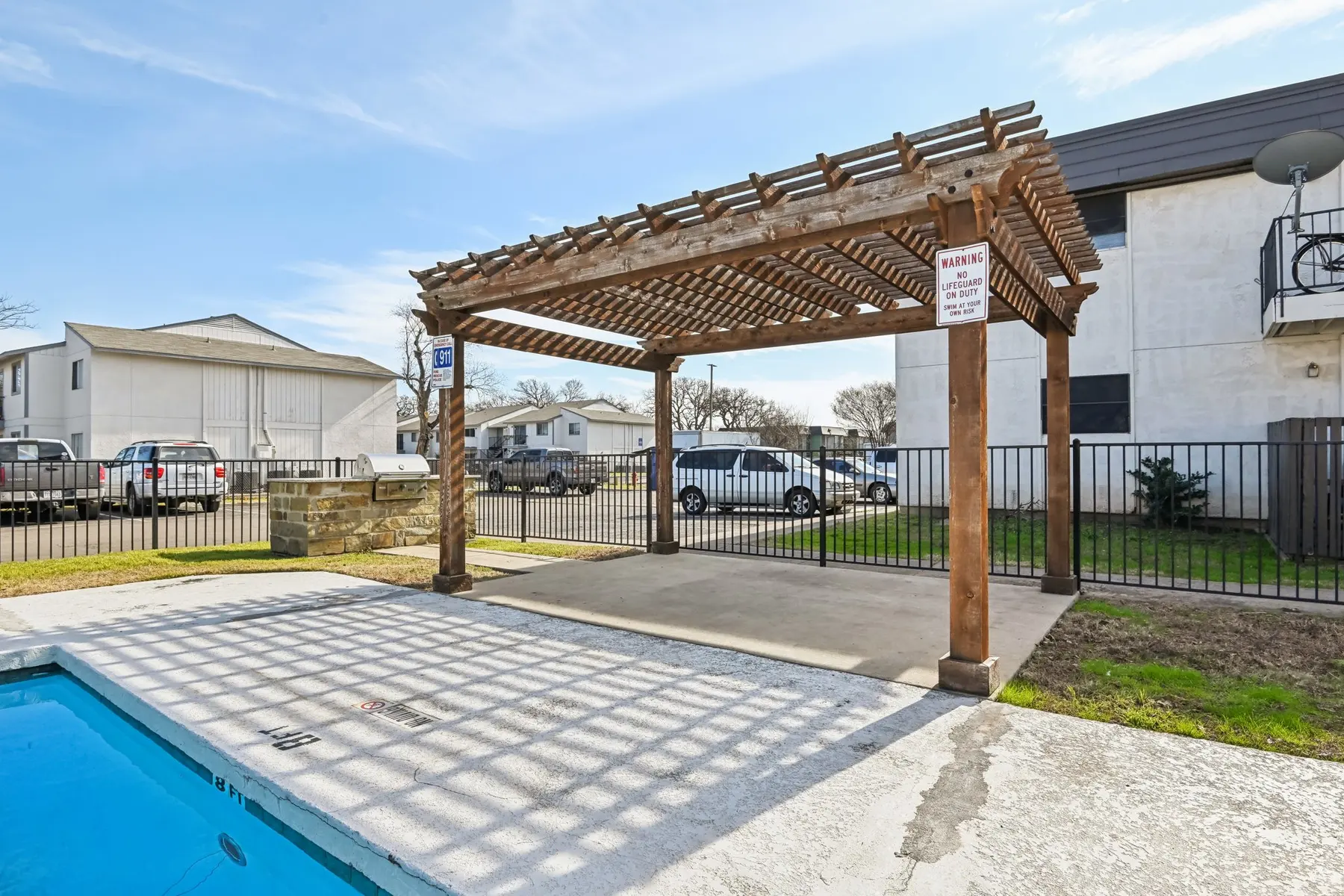 A poolside area featuring a wooden pergola and a swimming pool. The pergola is surrounded by a green lawn and there are parked cars in the background. A sign is attached to the pergola, indicating pool rules. The sky is clear and blue.