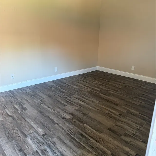 An empty room with wooden flooring and light-colored walls, featuring a small baseboard. There are no furniture or decorations present in the space.