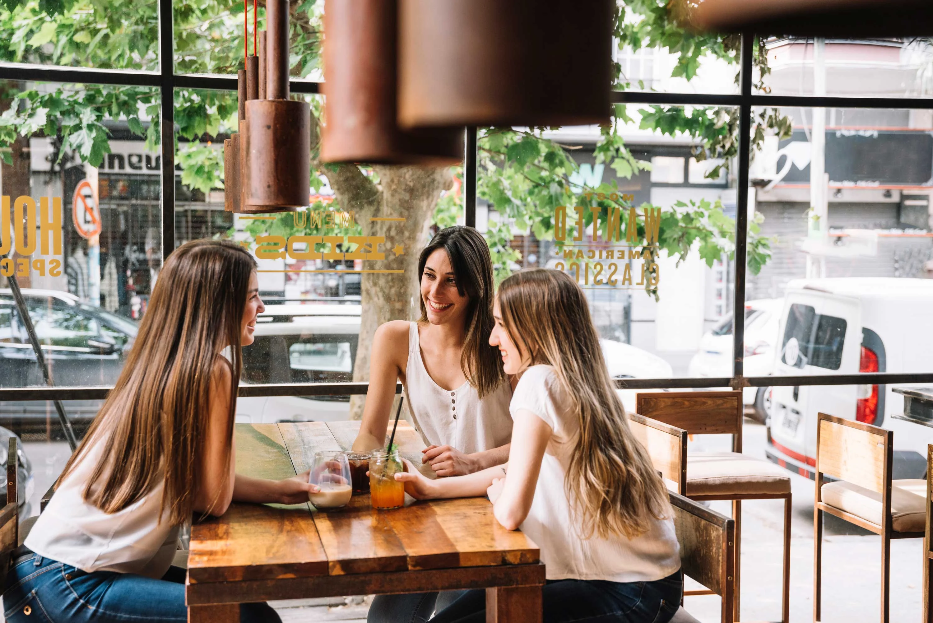 Friends Gathering at a Cafe Three young women chatting at a wooden table in a cafe with large windows and greenery outside.