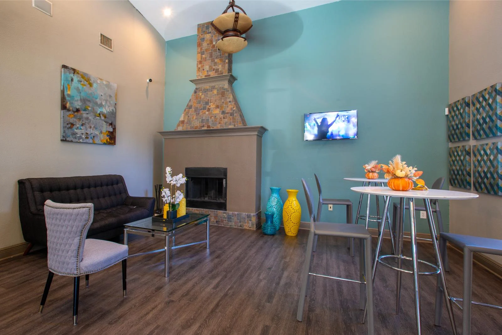 A stylish modern living space featuring a black sofa, a decorative fireplace, a dining table with orange and yellow decorative items, and vibrant vases. A flat screen TV is mounted on the wall, and the space has a cozy ambiance with wooden flooring.