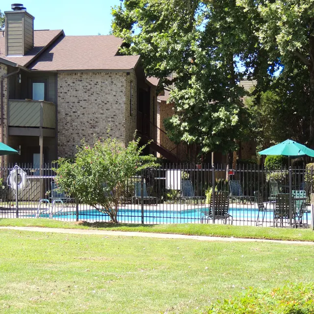 View of a swimming pool area surrounded by a fence, with lounge chairs and umbrellas, next to an apartment building.