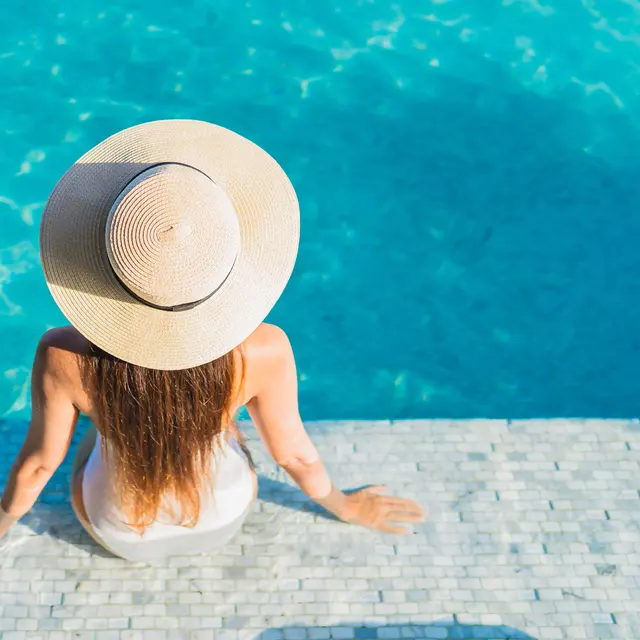 A person sitting at the edge of a pool, wearing a large sunhat and a white swimsuit, gazing at the water.