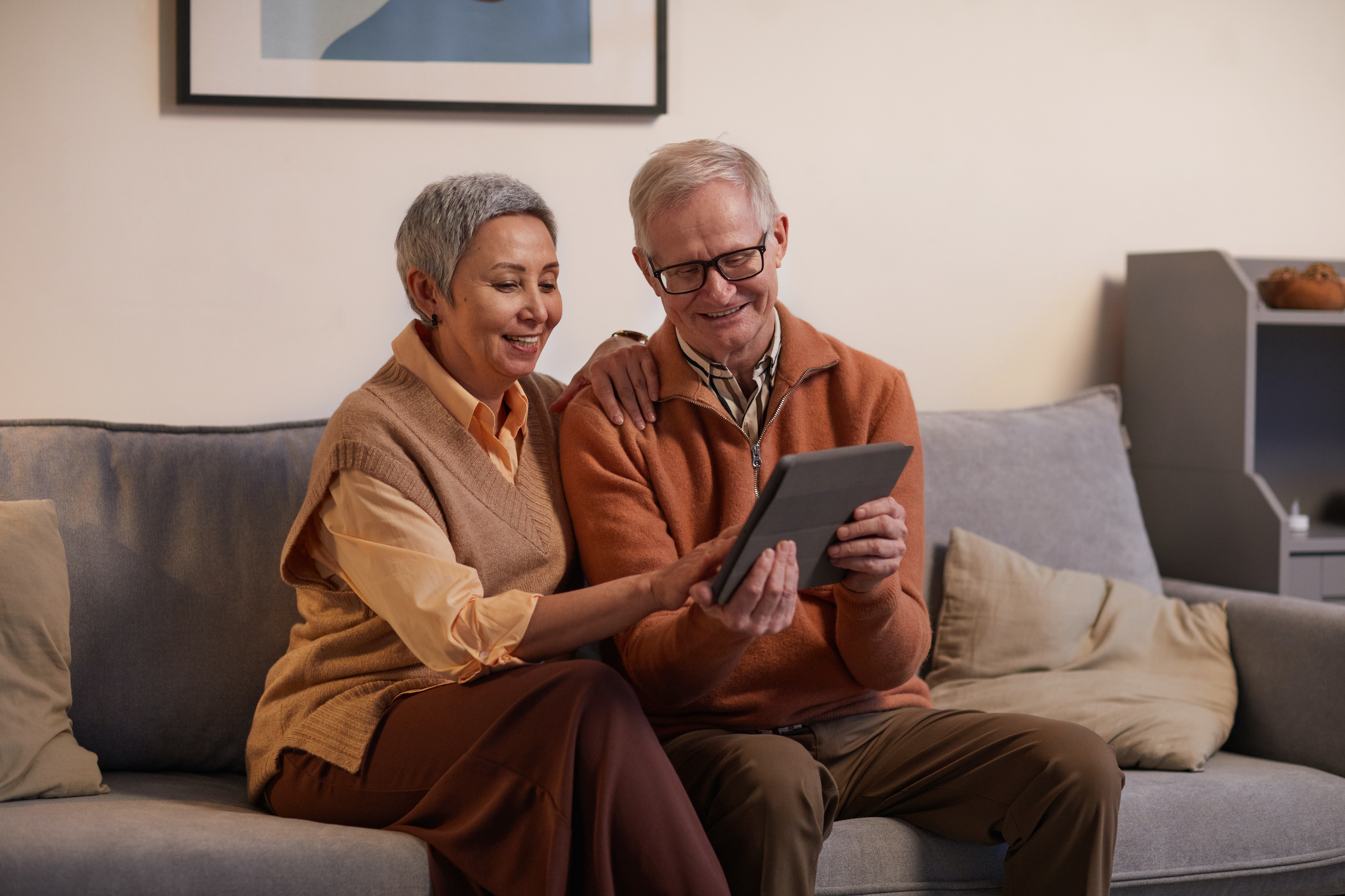 An elderly couple sitting together on a sofa, looking at a tablet and smiling. The woman has short gray hair and is wearing a light sweater, while the man has glasses and is dressed in an orange sweater. There's a cozy living room setting with light-colored decor.
