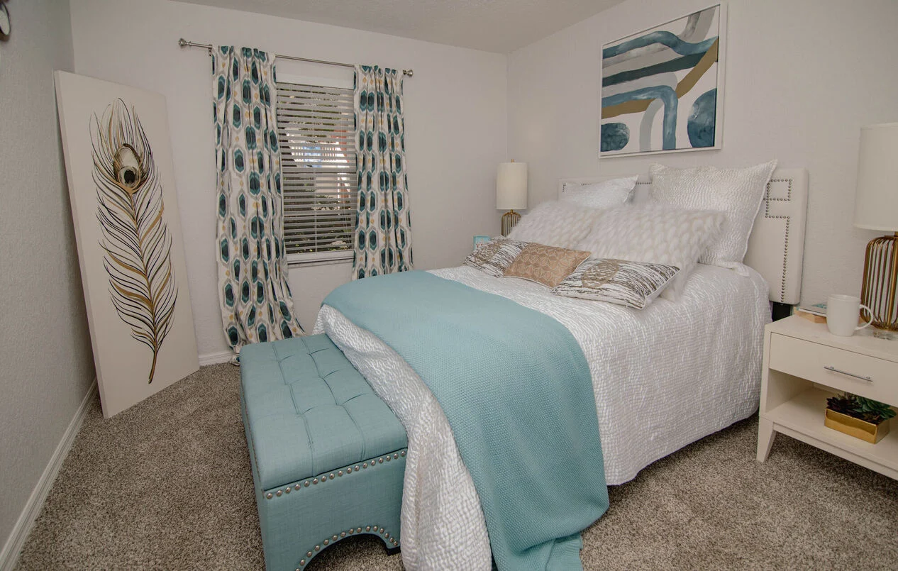 A cozy bedroom featuring a large bed with white bedding, a turquoise throw, and decorative pillows. There is a teal ottoman at the foot of the bed, and the walls have light-colored paint with a peacock feather artwork and sheer curtains with a geometric design.