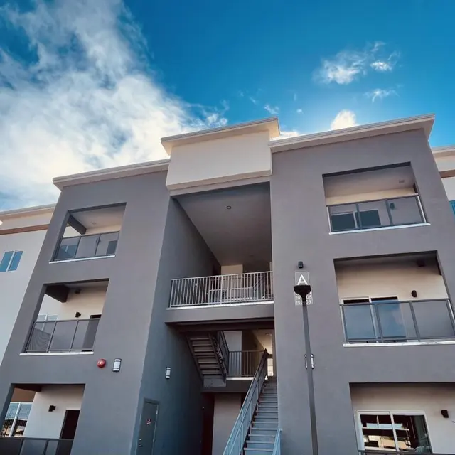 A modern multi-story apartment building with a mix of gray and white exterior, featuring balconies, large windows, and outdoor stairs leading to the entrance.