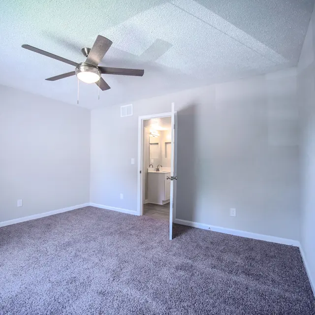 A spacious empty room with light gray walls, a ceiling fan, and a carpeted floor. A door to a bathroom is visible in the background.