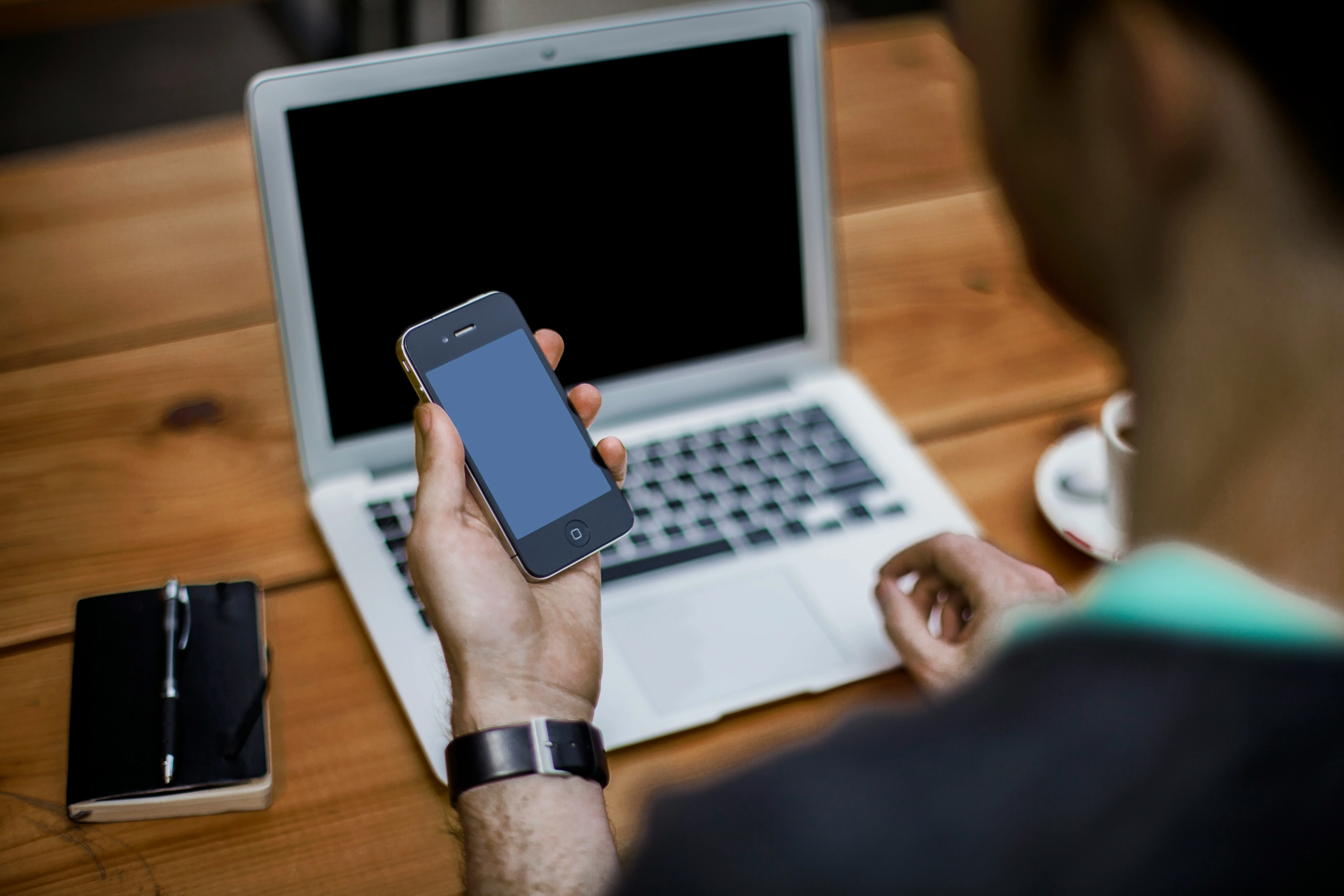 A person holding a smartphone in one hand, looking at it, with a laptop open in front on a wooden table. There is also a notebook and pen beside the laptop.