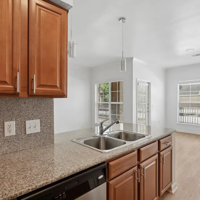 View of a modern kitchen with wooden cabinets, granite countertops, and a double sink overlooking a living area with large windows.