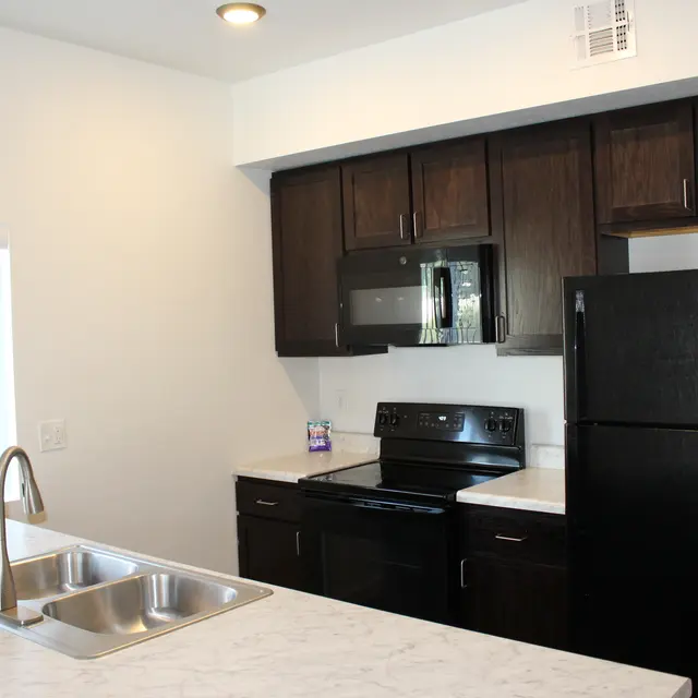 Stylish Modern Kitchen A modern kitchen featuring dark wooden cabinets, a black refrigerator, and an electric stove. The kitchen island has a double sink and a marble countertop with a light-colored finish. Natural light comes in from a window with vertical blinds.