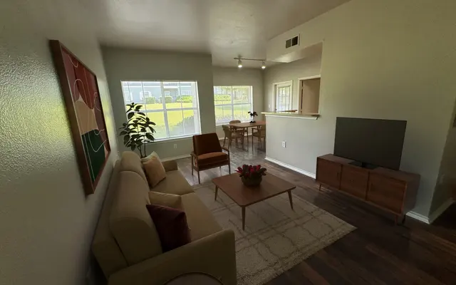 A cozy living room featuring a beige sofa, an armchair, a coffee table with a flower vase, and a TV stand with a television. Natural light pours in through large windows with views of greenery outside.