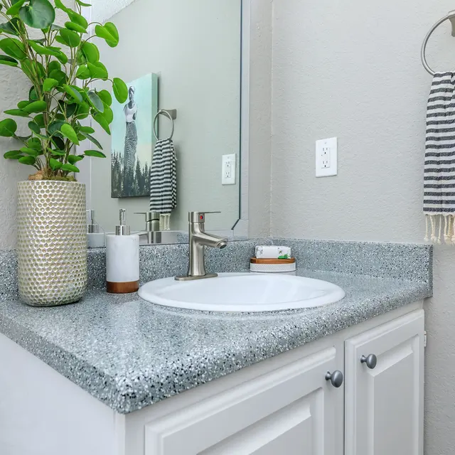 A modern bathroom sink area featuring a speckled gray countertop, a round sink, and a sleek faucet. A decorative plant is in a vase next to the sink. A striped towel hangs on a ring nearby and a small artwork is mounted on the wall.