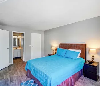 A cozy bedroom featuring a queen-size bed with blue bedding and two lamps on bedside tables. A bathroom is visible through a doorway. The walls are painted gray and the floor is covered with wooden tiles.