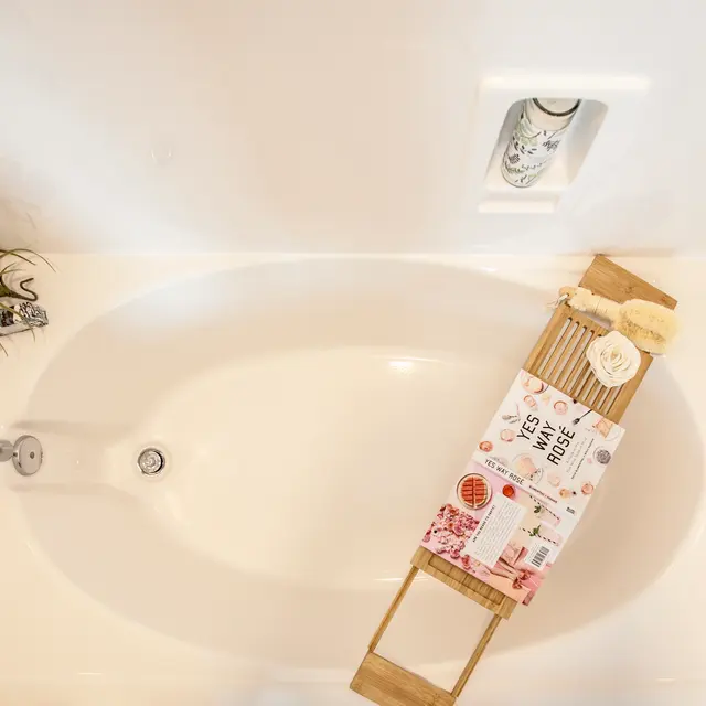 An overhead view of a clean, white bathtub with a minimalist design. There is a decorative wooden tray with various items placed on it, including a small potted plant and an assortment of bath products. The bathtub is empty and has a modern faucet visible.