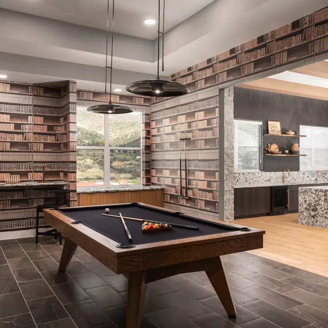 A stylish game room featuring a pool table in the center, surrounded by wallpaper that mimics bookshelves. The room has modern lighting and large windows showing a scenic view outside.