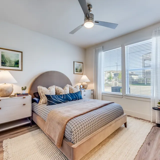 A modern and cozy bedroom with a large bed, light-colored walls, and two windows letting in natural light. The room features decorative lamps, a soft rug, and stylish furniture.