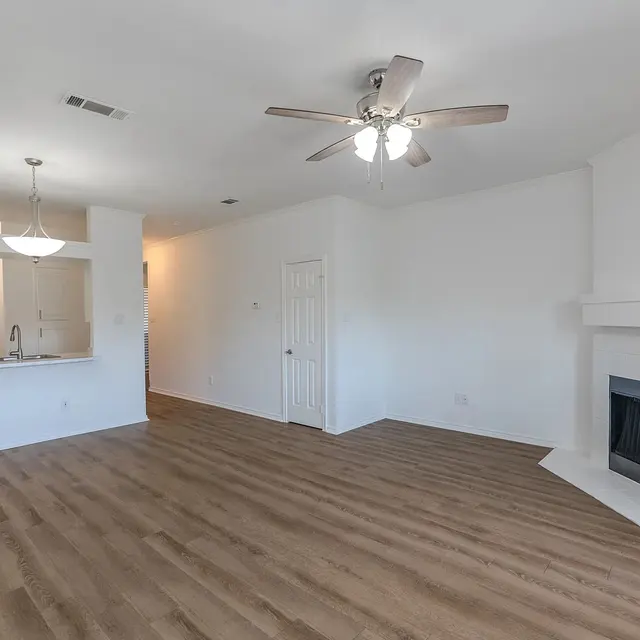 A bright and spacious living room featuring wooden flooring, a ceiling fan, and a fireplace. The room has white walls and an open layout that connects to a kitchen area in the background.