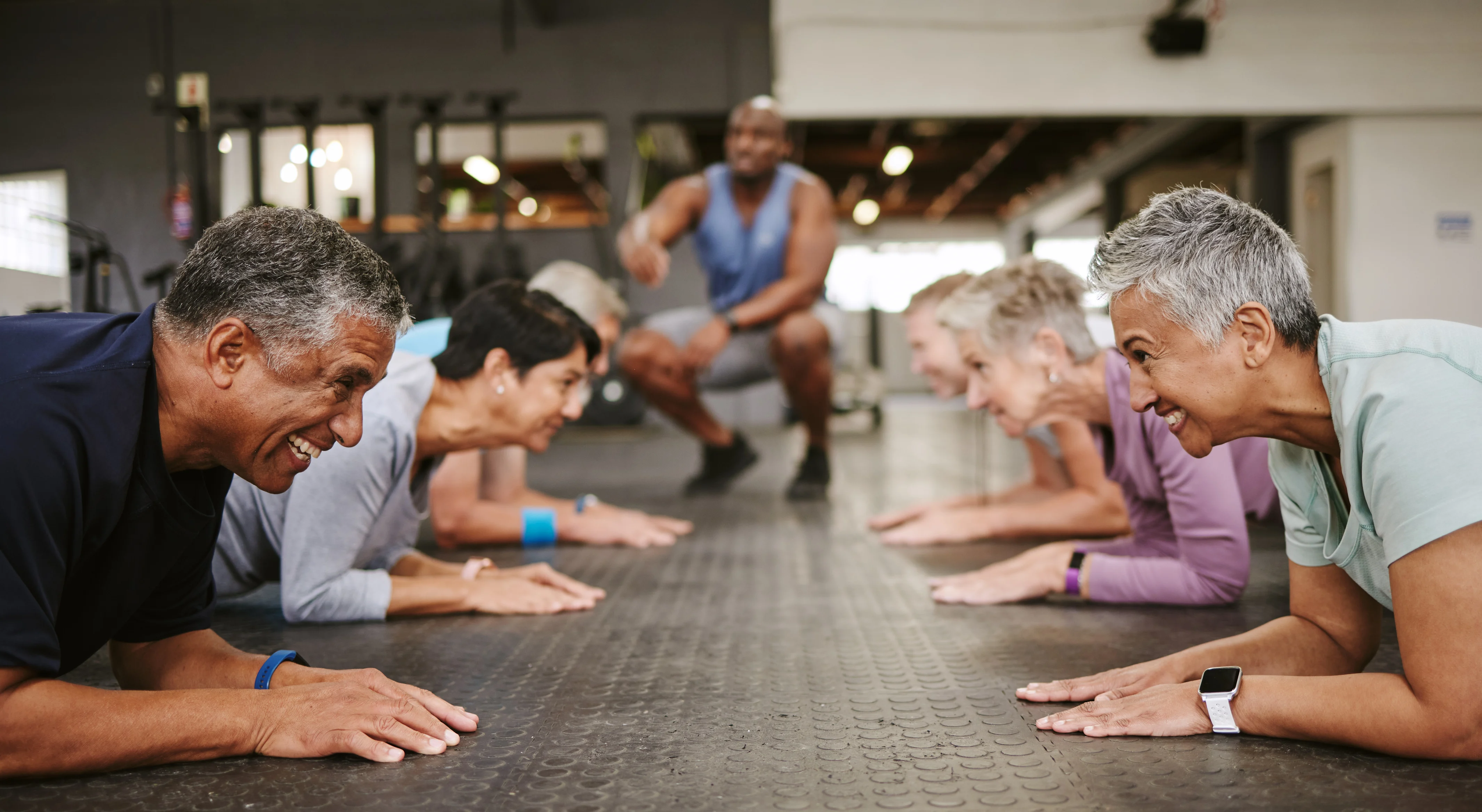 A diverse group of older adults engaged in a plank exercise in a gym, while a trainer encourages them. Everyone appears focused and determined.