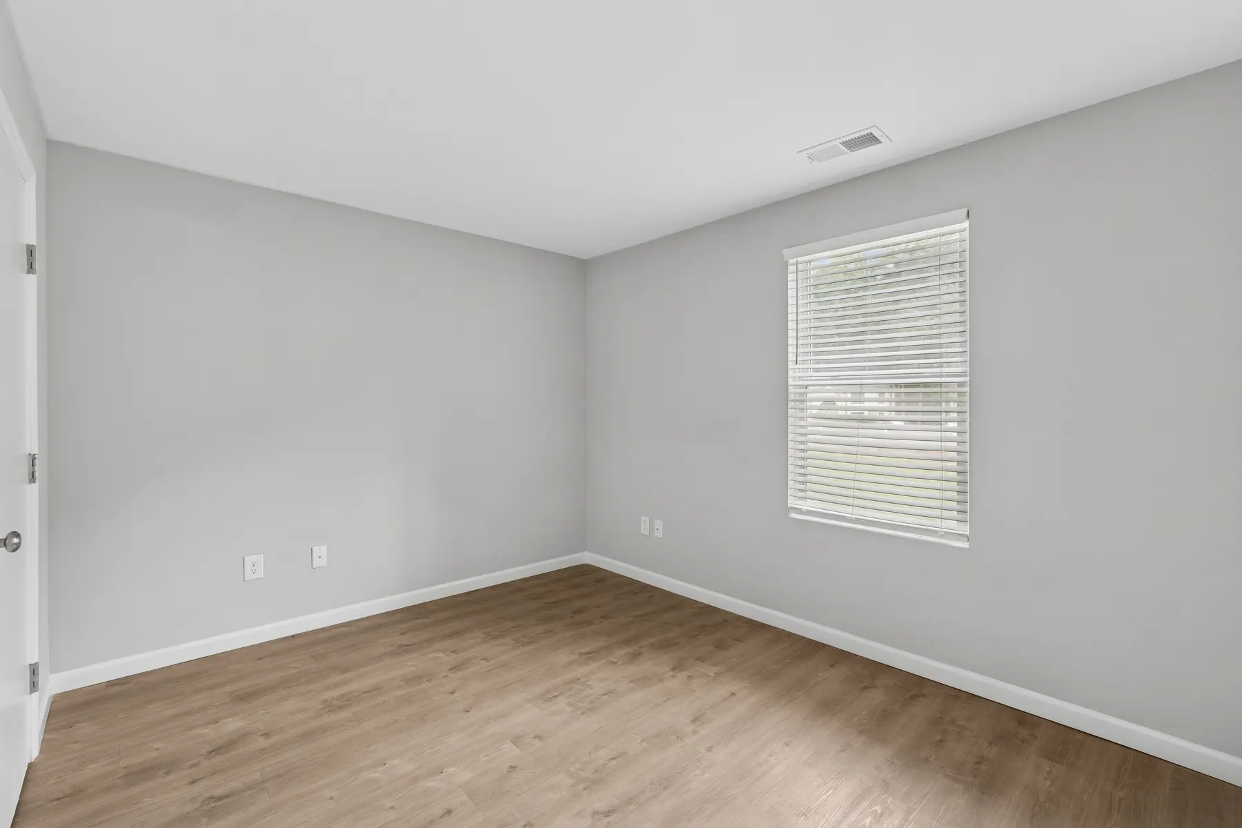 An empty room with light gray walls and a light brown wooden floor. There is one window with blinds on the right side.