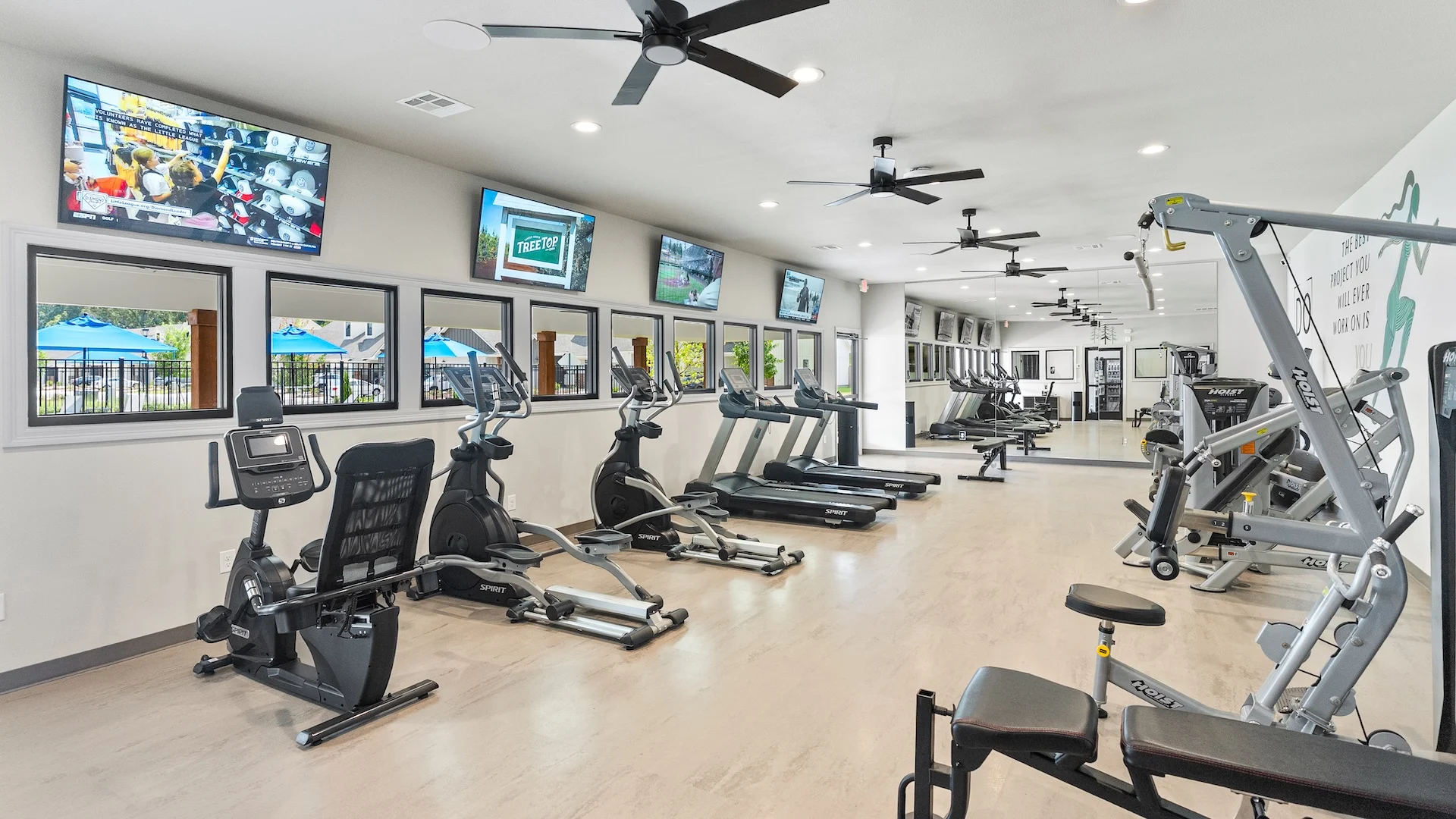 Interior of a Modern Gym A bright and modern gym interior featuring various workout equipment including treadmills, ellipticals, and weight machines. Multiple flat-screen TVs are mounted on the walls and there are large windows showing a view of an outdoor pool area.