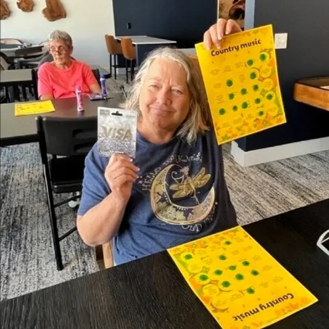 A woman with long hair smiling and holding a Visa gift card in front of a yellow bingo sheet for country music. In the background, another woman is seated at a table, also smiling.