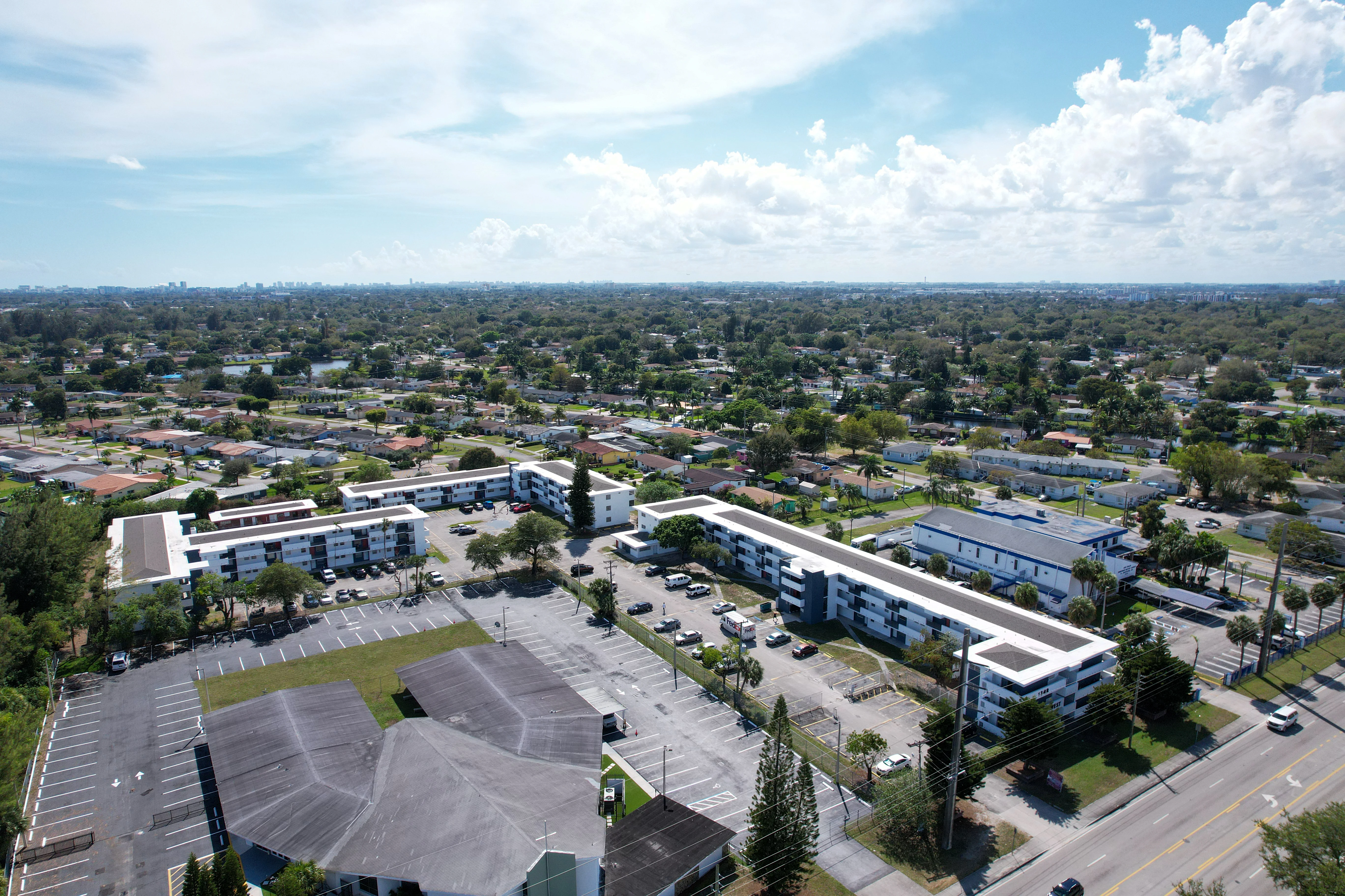 Aerial view of a suburban area featuring a residential complex surrounded by green spaces and roads under a clear blue sky with scattered clouds.