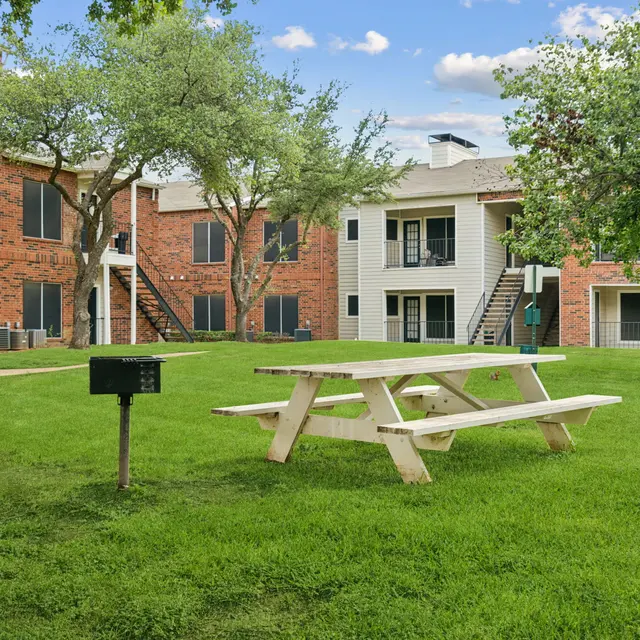 A well-maintained apartment complex courtyard featuring green lawn, picnic table, and trees with an exterior view of the buildings.