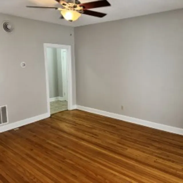A spacious living room with hardwood flooring, a ceiling fan, and gray walls. A doorway leads to another room, and a kitchen can be seen in the background.