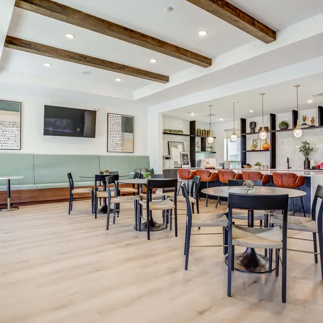 A modern lounge area featuring wooden beams, round tables with chairs, a sectional couch, and a bar counter in a bright interior.