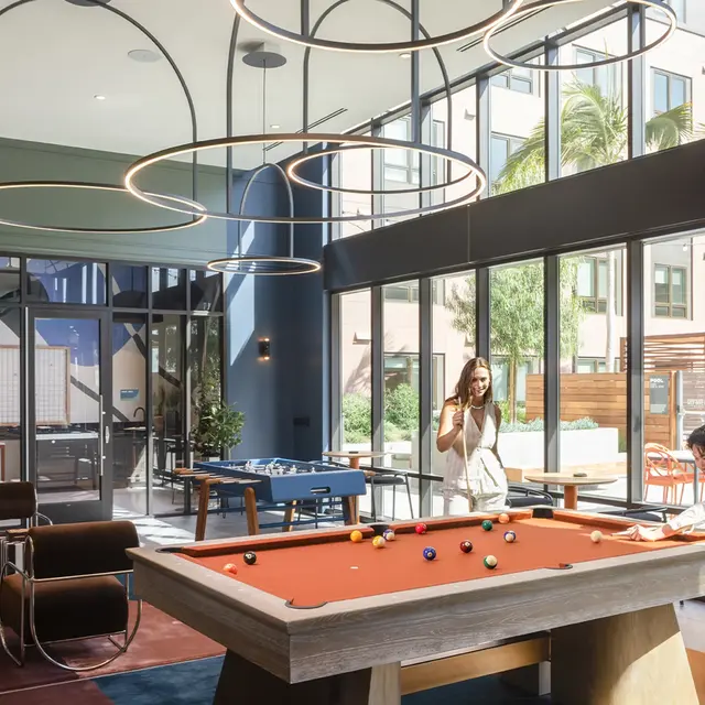A modern recreational room featuring a billiards table with an orange felt top, pool cues, and colorful decor. There are large windows allowing natural light, and abstract lighting fixtures hanging from the ceiling. Two people are engaged in activities related to the game, with one taking a photo while the other plays billiards.